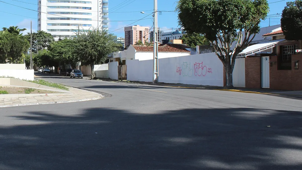 Rua de Campina Grande, na Paraíba, ao meio-dia: sombra recortada de árvore no asfalto, prédios e casas sob céu azul.
