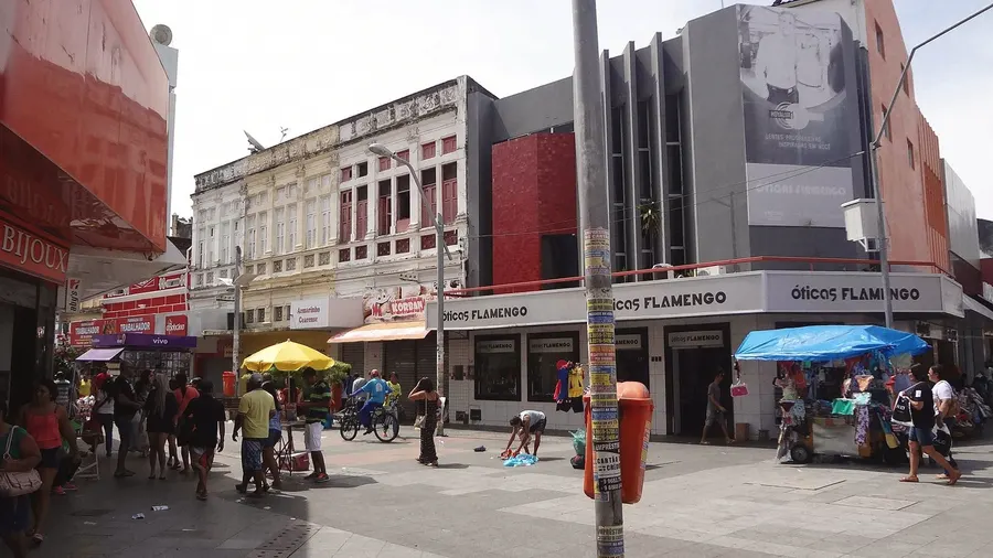 Rua do Comércio, no centro de Maceió, ao meio-dia: lojas abertas, guarda-sol de ambulante e gente no calçadão.